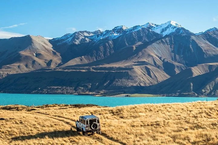 Ein 4WD-Fahrzeug fährt auf einer Schotterpiste auf einen türkisfarbenen See und schneebedeckte Berge zu.